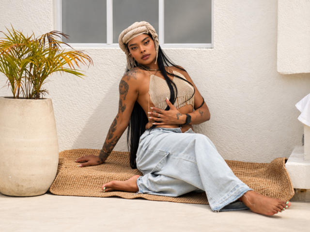 EmmaEbony with tan skin, black hair, tattoos, beige tank top, denim skirt, beige beanie, sitting on burlap, hands on tits, bare feet, potted plant, white wall, window.