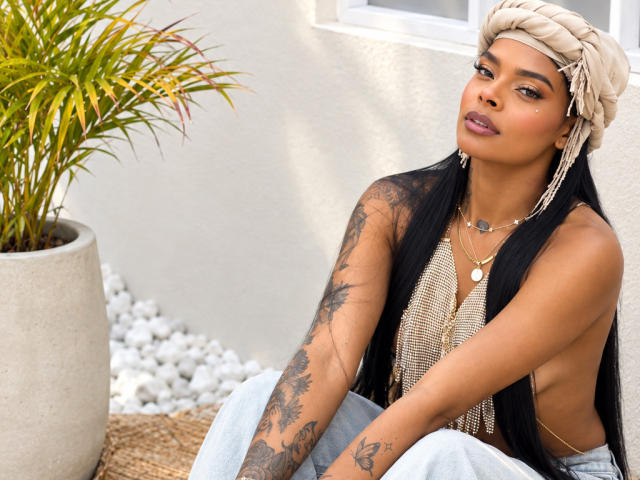 EmmaEbony with long black hair, tan skin, tattoos on arm and hand, wearing a beige headwrap, gold necklace, and a sleeveless top, sitting next to a potted plant.
