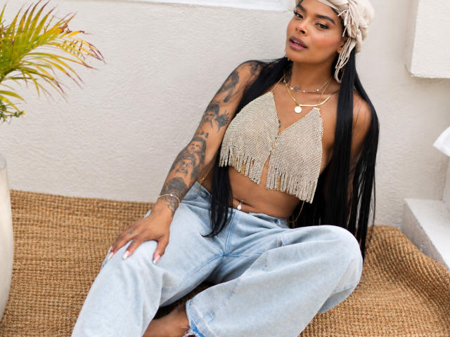 EmmaEbony with long black hair, wearing a beaded top with fringes, light blue jeans, and a white knitted hat, sitting on a woven rug, showing tattoos on her arm.