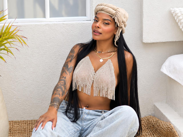 EmmaEbony with long black hair, tan skin, tattooed arm, beige headwrap, fringe top, showing small boobs, sitting, wearing light jeans, gold necklace, and belly button piercing.