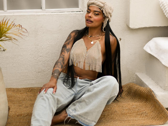EmmaEbony with long black hair, beige headwrap, fringe crop top, tattoos on arm, light jeans, sitting on burlap, looking at camera, wearing necklaces, brown skin, confident, sexy.