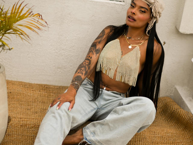 EmmaEbony with long black hair, wearing a fringed beige top, tattoos on her arm, light blue jeans, and jewelry, sitting on a burlap-covered surface, looking at the camera.