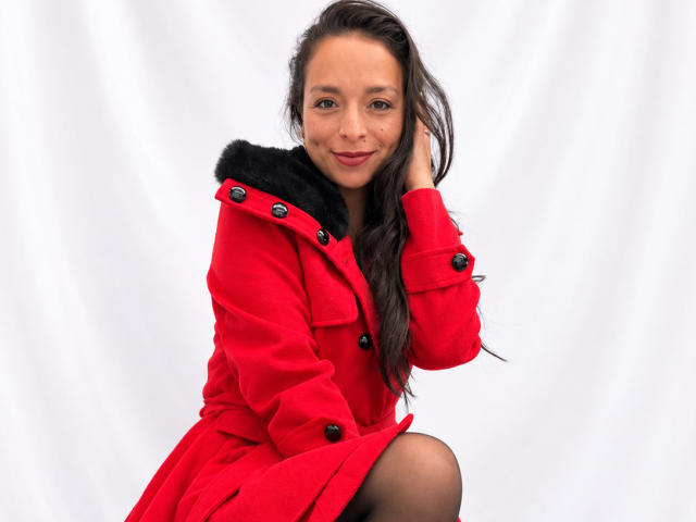 StangelarRivera, red coat with black fur collar, sitting, smiling, dark hair, one hand in hair, black pantyhose, white background, cute, attractive, medium tits.