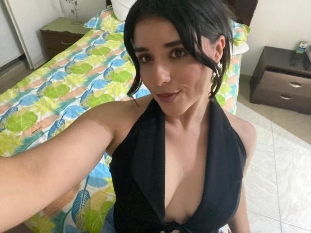 VickyMegan with black hair in pigtails, wearing a black halter top showing her cleavage, taking a selfie, her brown eyes and slight smile visible. Boobs are partially exposed.