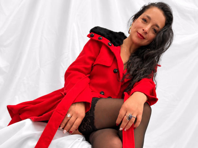 StangelarRivera with long black hair, red coat, black lace stockings, and ringed fingers, sitting on white cloth, showing black lace top and thigh.