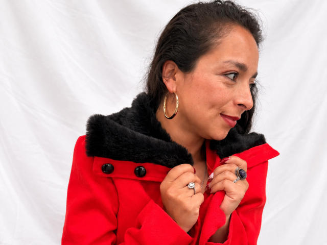 StangelarRivera with medium brown skin, black hair, and red coat with black fur collar, holding it with hands showing rings and blue gemstone, wearing gold hoop earrings.