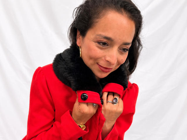 StangelarRivera with medium tits, wearing a red coat with black fur collar, holding buttons, smiling slightly, brown eyes, dark hair, gold hoop earrings, ring, white background.