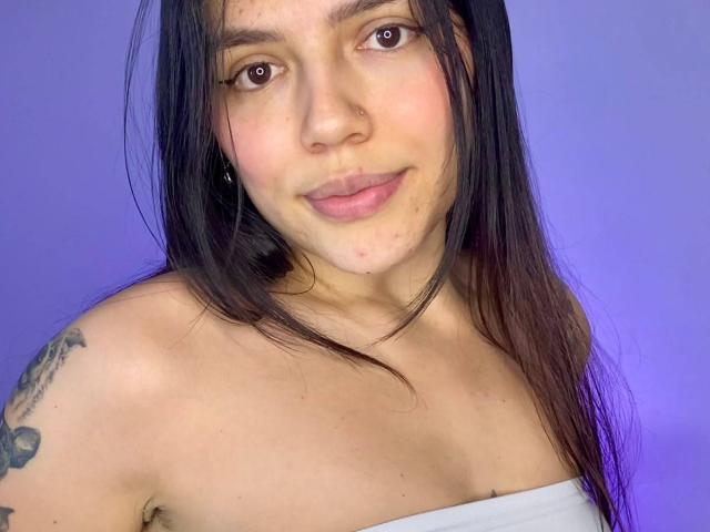 SophiaMarx with long black hair, light skin, small nose ring, subtle smile, bare shoulders, white strap, tattoo on left arm, purple background, soft light, gentle expression.