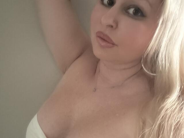 SeleneMoi has blonde hair, pouty pink lips, and is looking up with sultry eyes, showing her bare tits and a small necklace, arm raised in a soft-lit selfie.