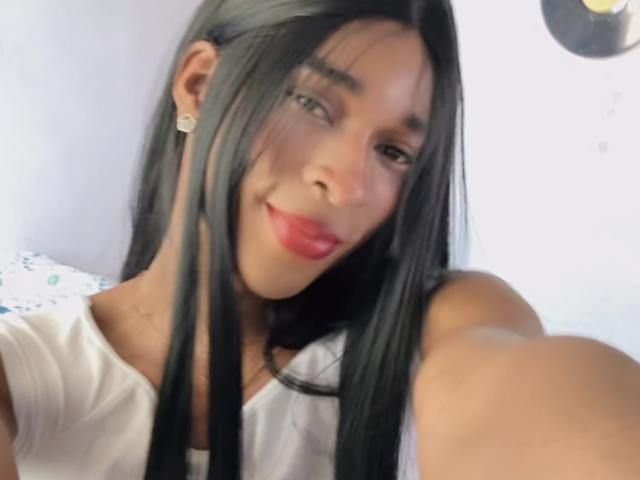 IlovetsCutie met lang zwart haar, rode lippenstift, een wit shirt en oorbellen, die glimlachend naar de camera kijkt en deze vasthoudt voor een selfie. Haar kleine borsten zijn gedeeltelijk zichtbaar..