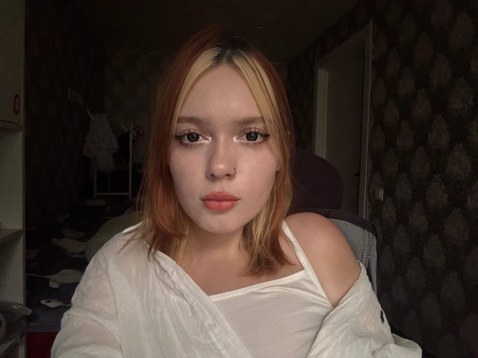Daniela Varon, com pele clara, cabelo loiro e laranja, lábios finos, camisa branca com um ombro descoberto, olhando para a câmera, sentada, em um quarto escuro, com papel de parede estampado, maquiagem discreta, pequeno piercing no nariz..
