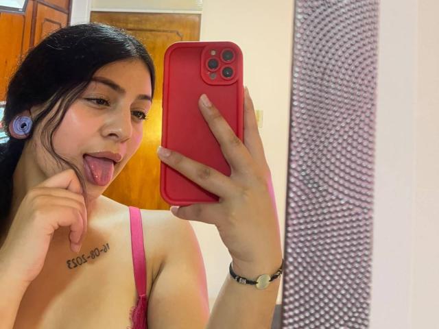 LonxEva with black hair, tongue out, wearing pink bra, holding red phone, small tits, tattoo on chest, purple earbuds, white wall, and textured right side in mirror selfie.