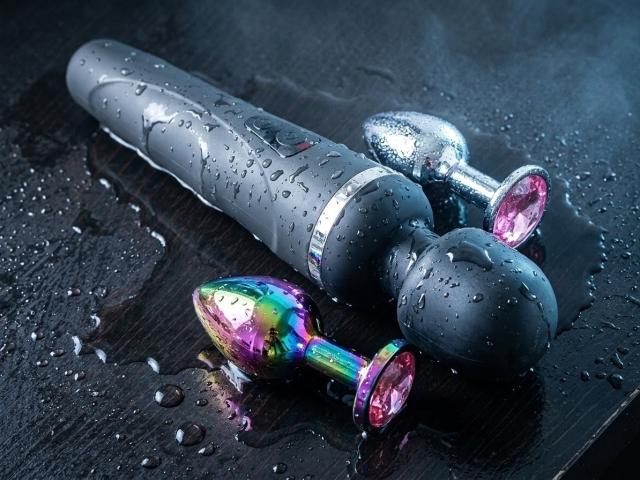 A wet, black vibrator with shiny, pink gem-like ends and a colorful, rainbow gem dildo on a dark, wet surface. Both are glistening with water droplets.