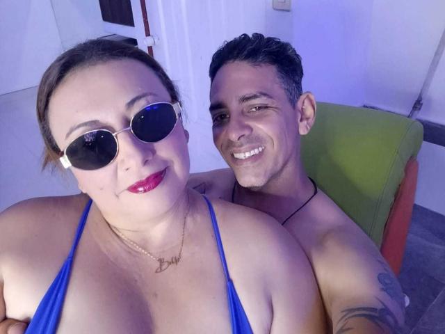 VictoriaSexOne with big tits, blue bikini top, and sunglasses, smiling next to a shirtless, tattooed guy with a happy smile, blue-tinted light, and green chair.