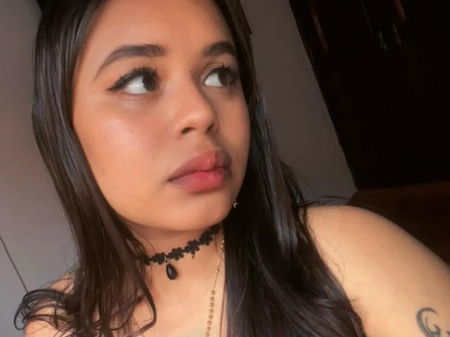 ChiaraBella with dark hair, looking to the side, has thick eyebrows, eyeliner, a black lace choker, and a small black pendant. Her lips are slightly parted.