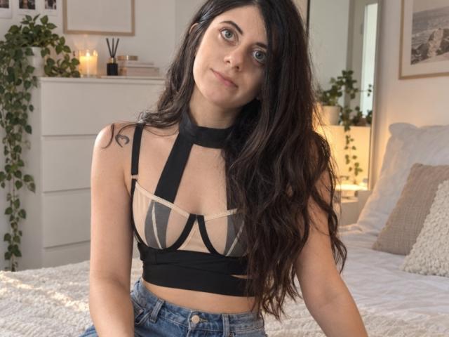 MilaSweets with long dark hair, wearing a black strappy top showing her tits and beige mesh, sitting on a bed in a cozy, lit-up room.