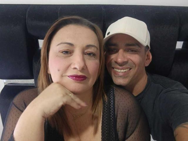 VictoriaSexOne with brown hair, pink lipstick, and black sheer top, has her chin on her hand, next to a smiling man in a white cap, black shirt, and earrings.