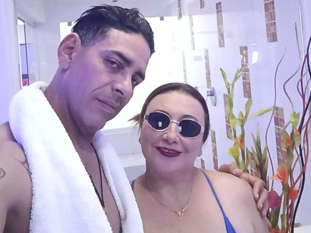 VictoriaSexOne with a towel around her neck, wearing blue straps, and round sunglasses, stands next to a man with short dark hair, smiling at the camera.