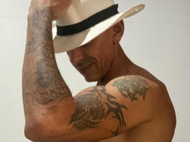 Tattooed VictoriaSexOne, shirtless with muscular arms, wearing a white hat with a black band, flexing, showing intricate tattoos on both arms.