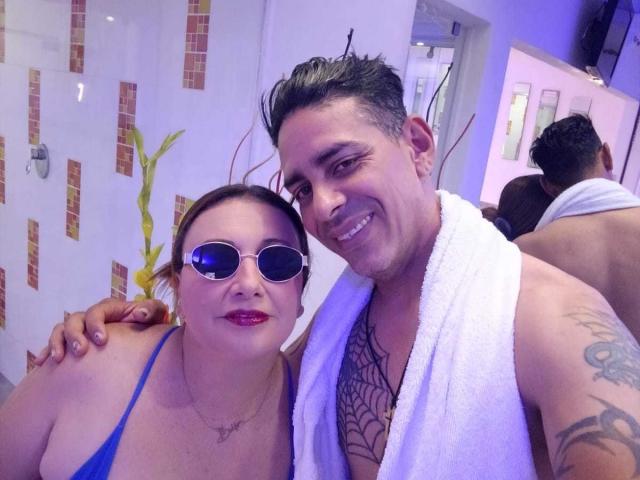 VictoriaSexOne with blue bikini top, small boobs, and sunglasses, standing next to a tattooed man with a white towel, smiling, in a brightly lit room with another shirtless guy.