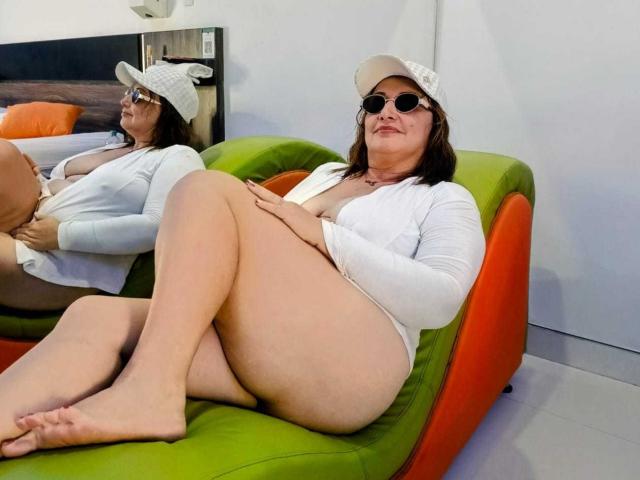 VictoriaSexOne with big ass, small tits, and a white cap, wearing sunglasses and a white cardigan, sitting on a green and orange chair, legs crossed, in a mirror reflection.