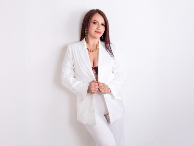CameronViviane with long brown hair, white blazer, white pants, gold necklace, and earrings, standing, smiling, hands on blazer, exposing her small, brown-tipped boobs.
