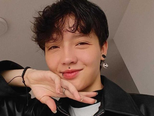LissyLuxe with short, messy black hair, a nose piercing, and a lip piercing, wearing a black leather jacket, smiling, and touching their chin with a ringed finger.