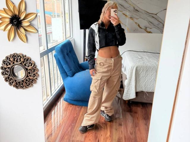 ElenaSaenz with pink hair, black crop top, tan cargo pants, and black shoes, taking a mirror selfie, showing her bare midriff and ass. Blue chair in the background.