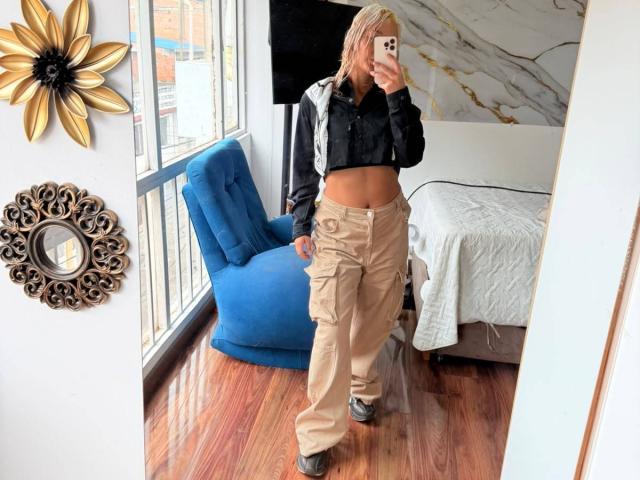ElenaSaenz with pink hair, black crop top, tan cargo pants, showing her midriff, taking a mirror selfie, standing on shiny wooden floor, in a modern room.