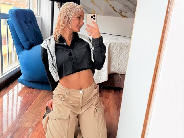 ElenaSaenz with wet blonde hair, black and white hoodie, tan cargo pants, showing her flat abs, taking a mirror selfie, holding a gold iPhone, in a bright room.