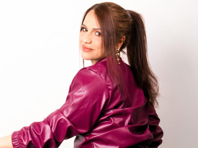 CameronViviane with long brown ponytail, wearing a shiny purple leather jacket, smiling at the camera, small gold earrings, white background. Boobs not visible, focus on upper body.