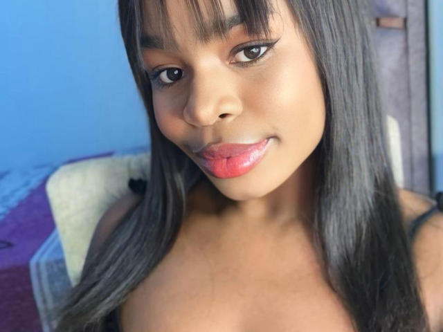 VenusWilly with dark straight hair, black eyeliner, and shiny pink lips, smiling with tongue slightly out, showing smooth brown skin, and bare shoulders.