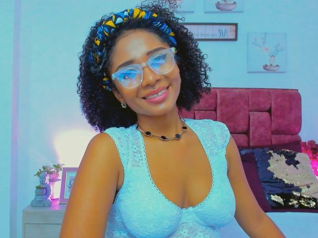 MeganWilliams with curly black hair, blue headband, clear glasses, smiling, wearing lace blue top, necklace, showing small tits, pinkish skin, cute, bedroom, pink headboard, nightstand.
