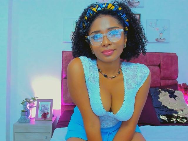 MeganWilliams with curly hair, blue headband, glasses, and a deep blue lace top showing her tits, sitting on a bed, smiling, wearing a necklace, in a brightly lit room.
