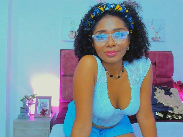 MeganWlliams with curly black hair, blue headband, glasses, and blue lace-up top, showing her tits, wearing blue shorts, smiling, in a brightly lit bedroom.