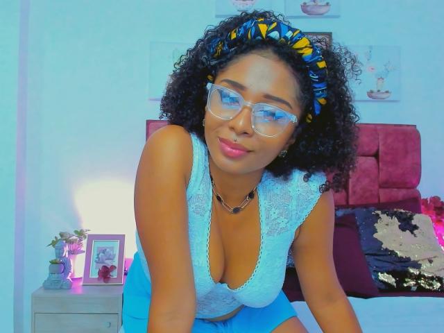 MeganWilliams with curly black hair, blue headband, glasses, white lace top, revealing cleavage, blue shorts, smiling, kneeling on bed, necklace, pink lips, bedroom, nightstand with photo frame.