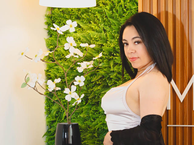 BellaFournierHot with black hair, white halter top, black sleeves down, holding back black sleeves, looking at camera, standing next to white flowers in black vase.