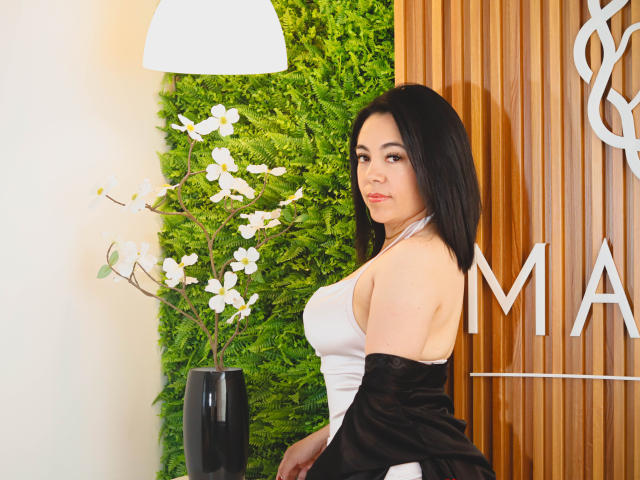 BellaFournierHot with black hair, white halter dress, black jacket off, showing her tits, standing next to white flowers in a black vase, wooden wall in background.