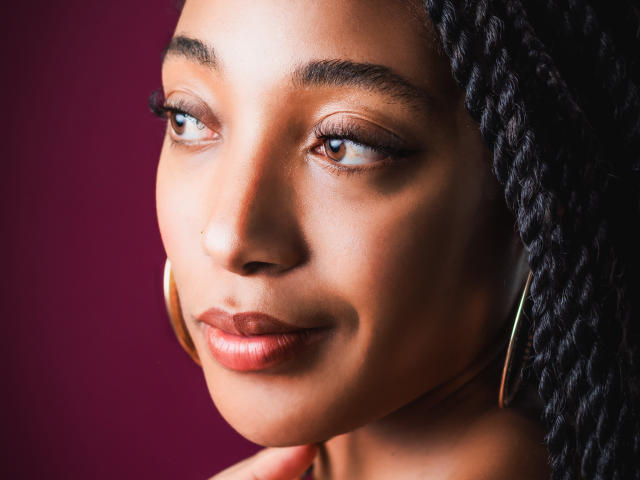 EsmeraldaRoe's face, dark brown skin, curly black hair, gold hoop earrings, subtle smile, brown eyes with long lashes, slight nose ring, glossy pink lips, looking to the side.