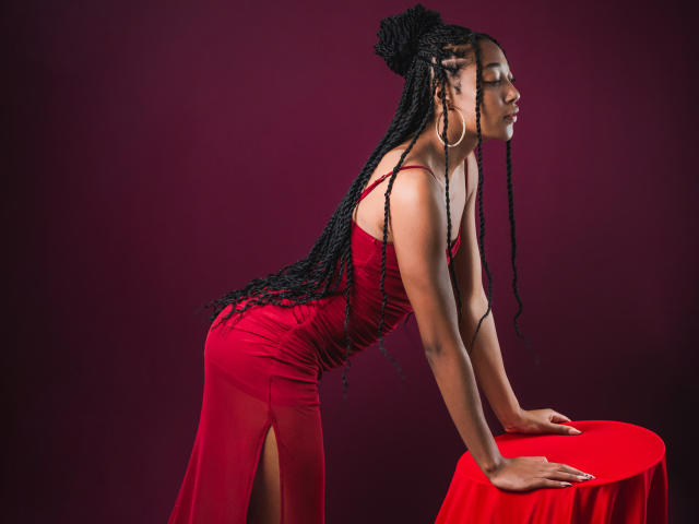 EsmeraldaRoe with long black braids, wearing a tight red dress, leaning on a red-covered stool, showing her ass, red background, looking to the side, hoop earrings.