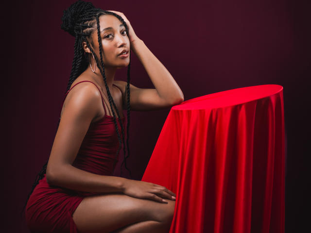 EsmeraldaRoe with dark braids, wearing a red dress, sitting by a red-covered table, hand on head, looking at camera, showing her ass and tits.
