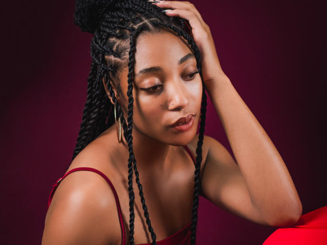 EsmeraldaRoe has dark braided hair, wearing a red dress, with her hand in her hair, looking down with a pout, and small gold hoop earrings against a dark background.