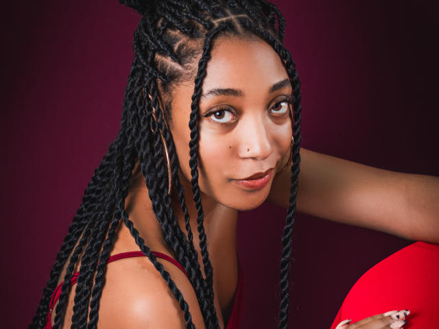 EsmeraldaRoe has long black braids, brown eyes, and a small nose piercing. She's wearing a red top and holding a red fan, with a serious expression.