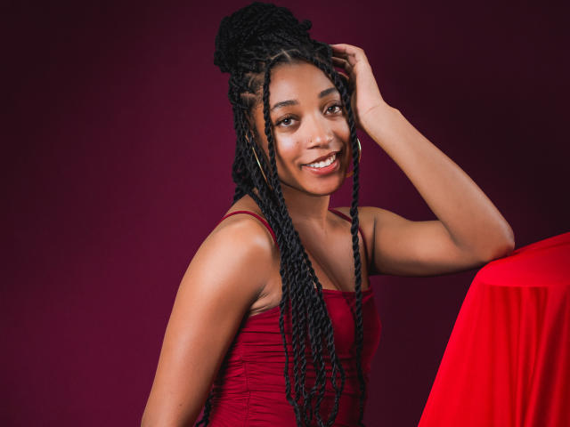 EsmeraldaRoe with long black braids, wearing a red dress, smiling at the camera, hand in hair, sitting next to a red-covered object. Her tits are small.