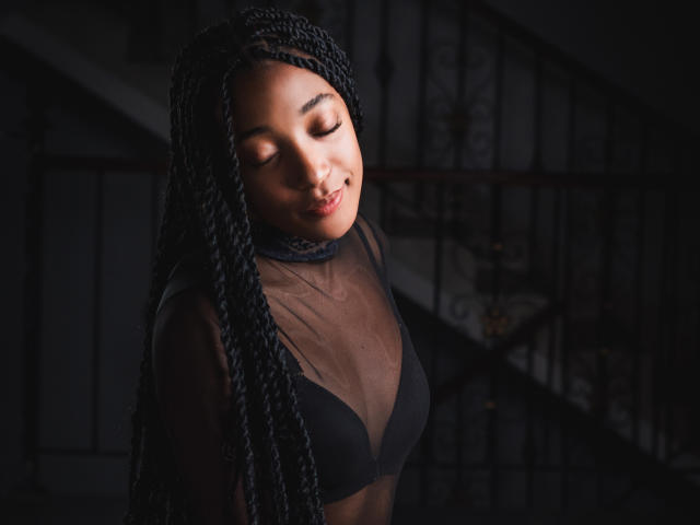 EsmeraldaRoe with dark braided hair, closed eyes, and a soft smile, wearing a sheer black top that reveals her small, perky tits and black bra. Dim, moody lighting.