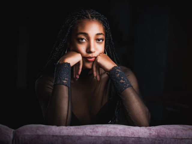 EsmeraldaRoe with braided hair, black lace sleeves, and hoop earrings, leaning on a pink couch, hands under chin, dark background, looking at camera with soft, sultry expression.