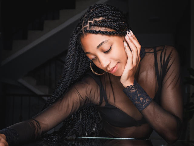 EsmeraldaRoe with long black braids, wearing a sheer black top, touching her cheek, smiling, showing her boobs, large hoop earrings, dark background, soft light, relaxed pose.
