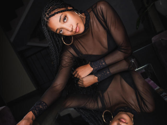 EsmeraldaRoe with long braids, wearing a sheer black dress, sitting on a glass table, arms crossed, large hoop earrings, smiling, looking up, dark room, reflective surface.