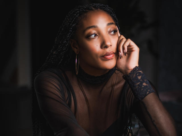EsmeraldaRoe has dark skin, braided hair, and is looking to the side with a slight smile, wearing a sheer black top, gold hoop earrings, and lace on her wrist.