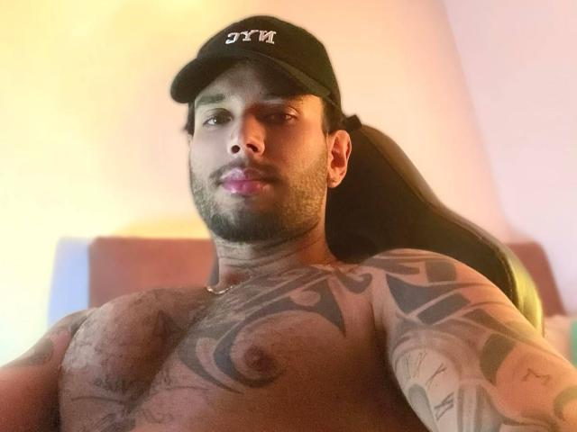 Aletargaryem, sin camisa y con tatuajes que cubren su pecho y brazos, lleva un gorro negro, tiene una coleta y barba, y mira hacia abajo con una ligera sonrisa..